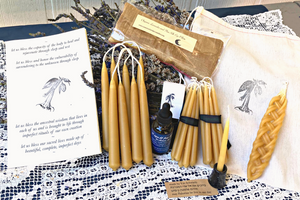 an offering of golden beeswax candles in different shapes and sizes alongside poetry offerings and herbs on a table