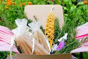 a summer deluxe heart of fire ritual box in the garden contains 12 pairs of shabbat candles, one braided Havdallah candle, a bottle of Molten Courage Oxymel, a bottle of Safe Passage Essence Blend and fresh sprigs of rosemary