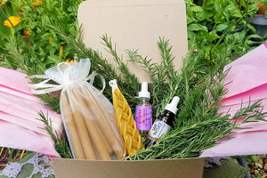 a heart of fire summer ritual box in the garden contains four pairs of shabbat candles, one braided Havdallah Candle, a bottle of Molten Courage, a bottle of Safe Passage and fresh sprigs of rosemary